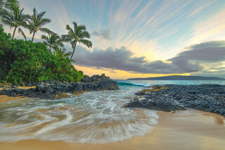 Palm trees frame a hidden golden sand cove at Secret Beach in Makena, Maui, during a glowing, warm tropical sunset. 