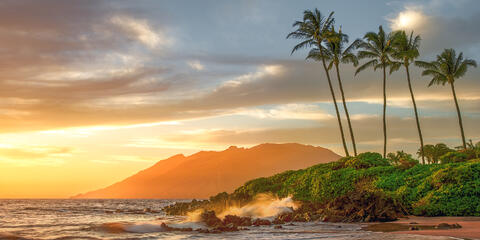 Golden sunset at Polo Beach in Maui with ocean waves, lava rocks, tall palm trees, and the West Maui Mountains in the background.