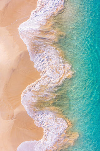 Top-down aerial view of turquoise waves and foamy white surf crashing onto the golden sand of Big Beach in Makena, Maui.