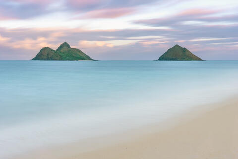Long-exposure view of the Na Mokulua twin islands from Lanikai Beach on Oahu under a soft pastel sunset sky.
