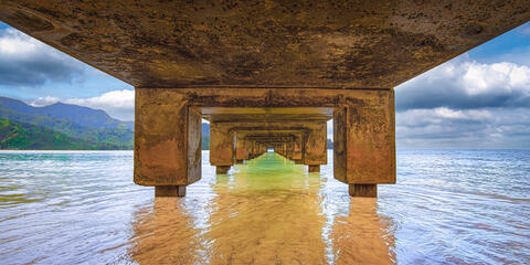 Symmetrical under-pier view of Hanalei Pier in Kauai showing concrete pillars framing clear golden water and green mountains.