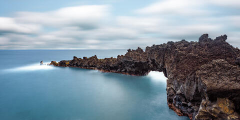 Long exposure photo of a natural black basalt lava archway over smooth blue ocean water at Wainapanapa State Park in Maui.