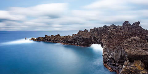 Long exposure photo of a natural black basalt lava archway over smooth blue ocean water at Wainapanapa State Park in Maui.