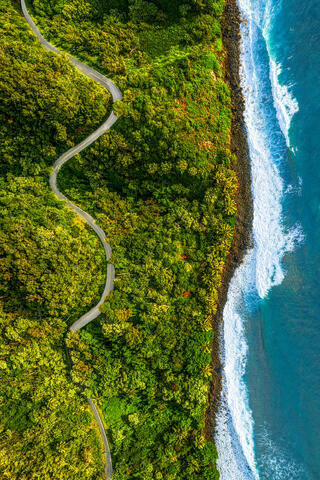 Top-down aerial view of the winding Hana Highway cutting through a lush green jungle alongside blue ocean waves crashing on Maui's shore.
