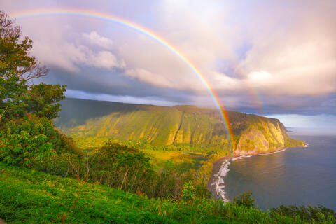 A vibrant double rainbow arches over the towering green cliffs and black sand beach of Waipio Valley at sunrise on Hawaii Island.