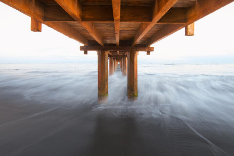Abstract long-exposure view beneath the wooden structure of Waimea Pier, showing weathered beams, pilings, and silky ocean water.