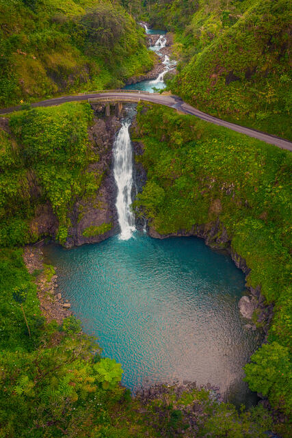 Aerial view of Makapipi Falls cascading under a bridge on the Road to Hana into a turquoise pool surrounded by lush green jungle in Maui.