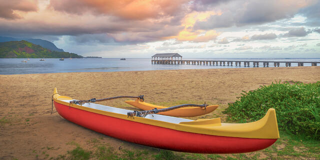 A red and yellow Hawaiian outrigger canoe rests on Hanalei Bay beach at dawn with Hanalei Pier and green mountains in Kauai.