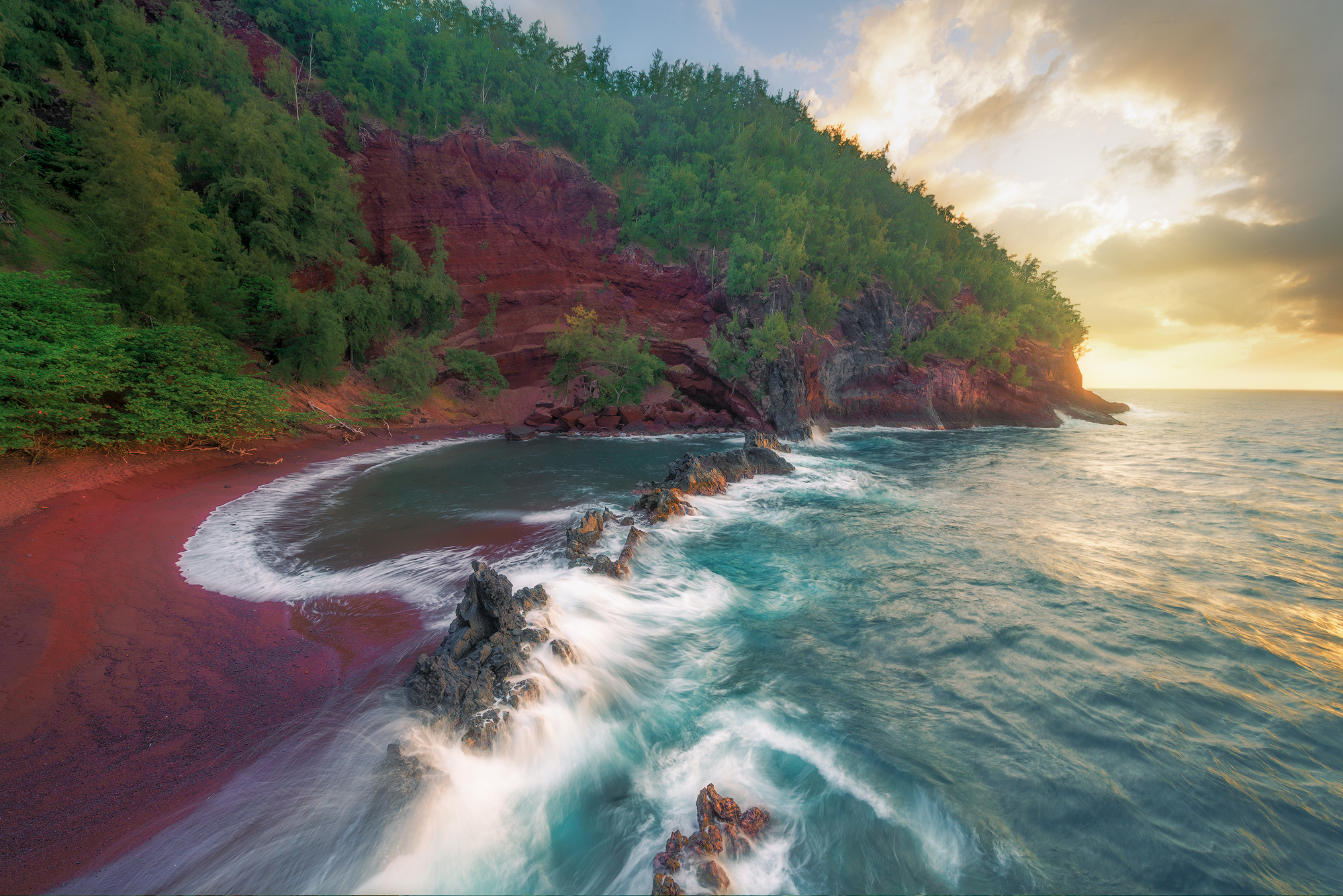 Kaihalulu Sunrise Sunrise over Kaihalulu Red Sand Beach in Hana, Maui, showing crimson cliffs, turquoise water, and a black lava rock barrier.