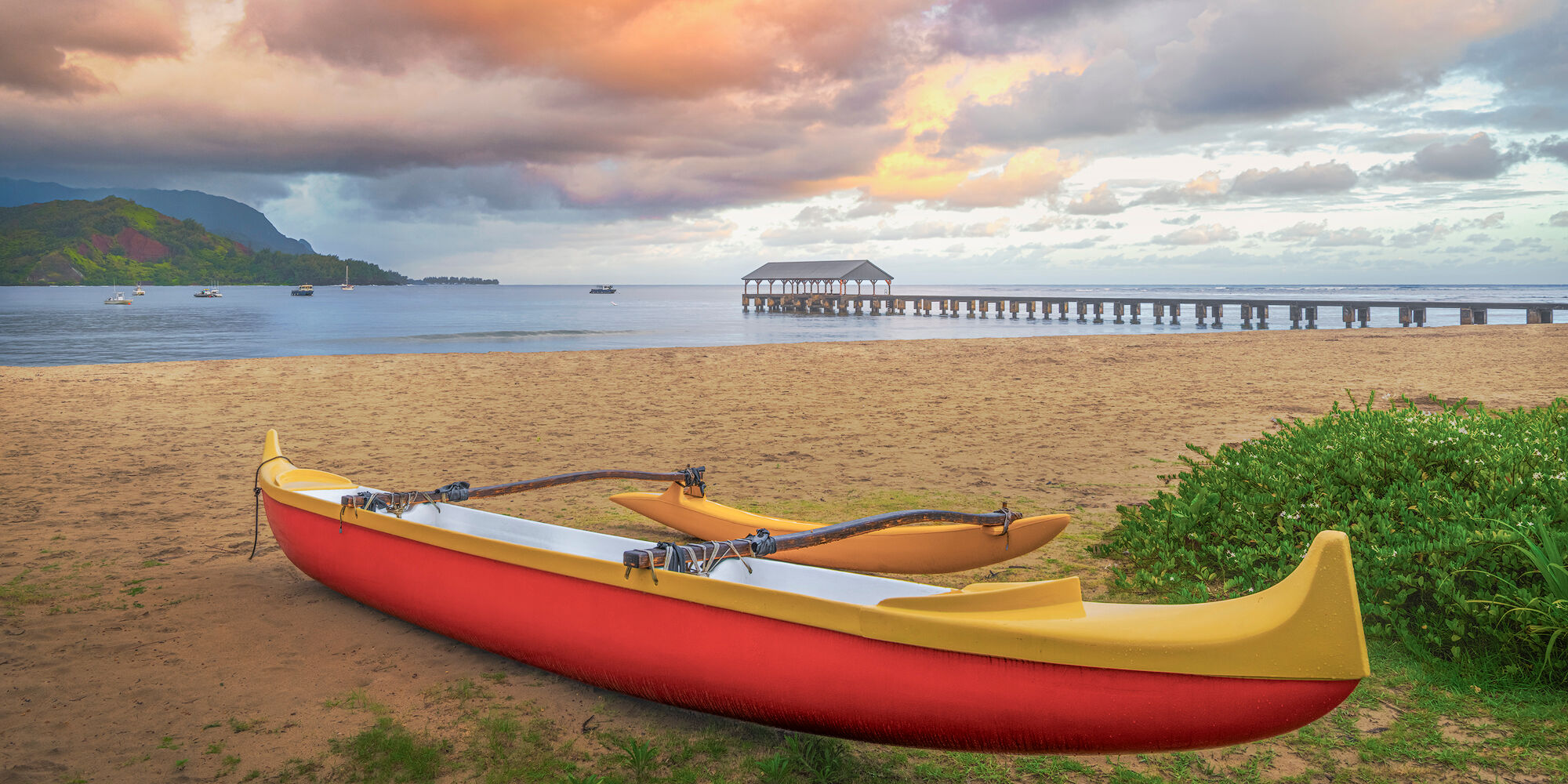 Outrigger at Hanalei Bay A red and yellow Hawaiian outrigger canoe rests on Hanalei Bay beach at dawn with Hanalei Pier and green mountains in Kauai.