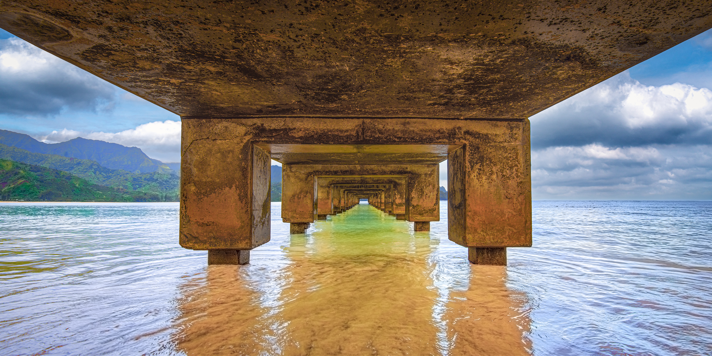 Tunnel Vision at Hanalei Symmetrical under-pier view of Hanalei Pier in Kauai showing concrete pillars framing clear golden water and green mountains.