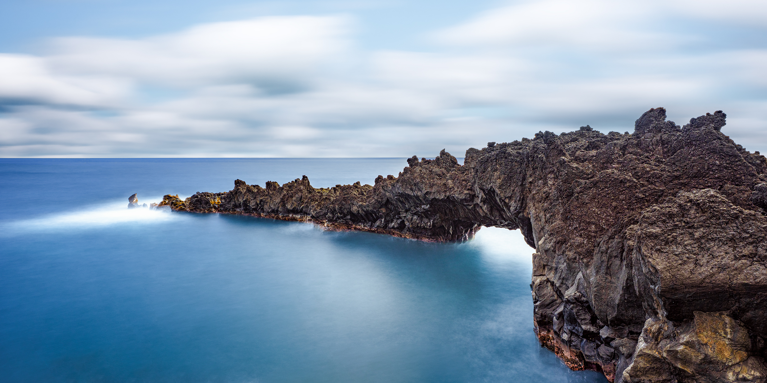 The Archway at Waiʻānapanapa Long exposure photo of a natural black basalt lava archway over smooth blue ocean water at Wainapanapa State Park in Maui.