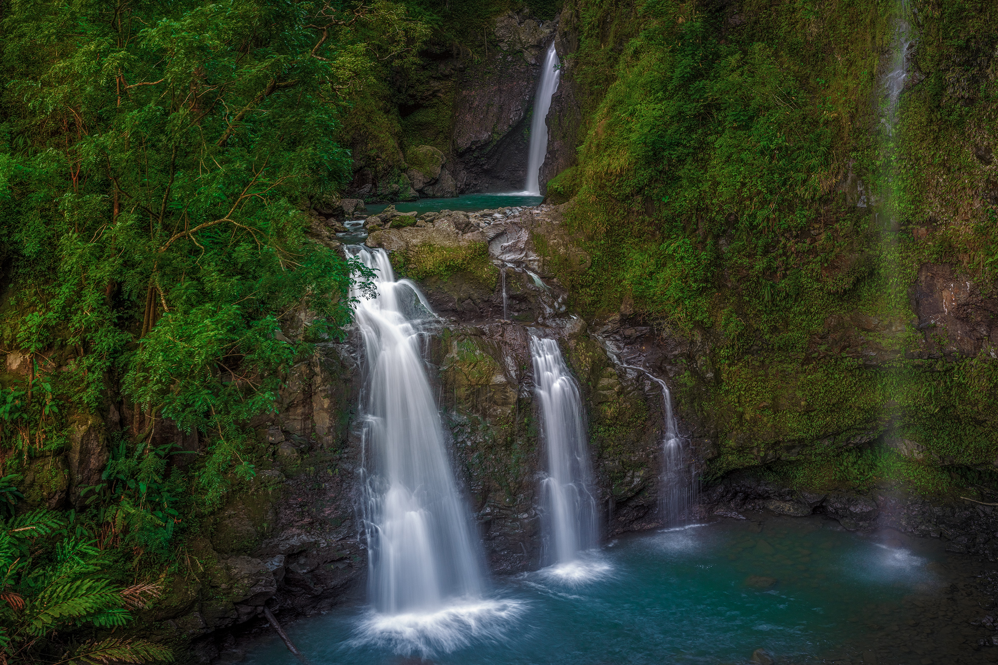Tiers of Waikani Long exposure of the multi-tiered Waikani Falls cascading over dark rocks into a turquoise pool surrounded by lush jungle on the Road to Hana in Maui.