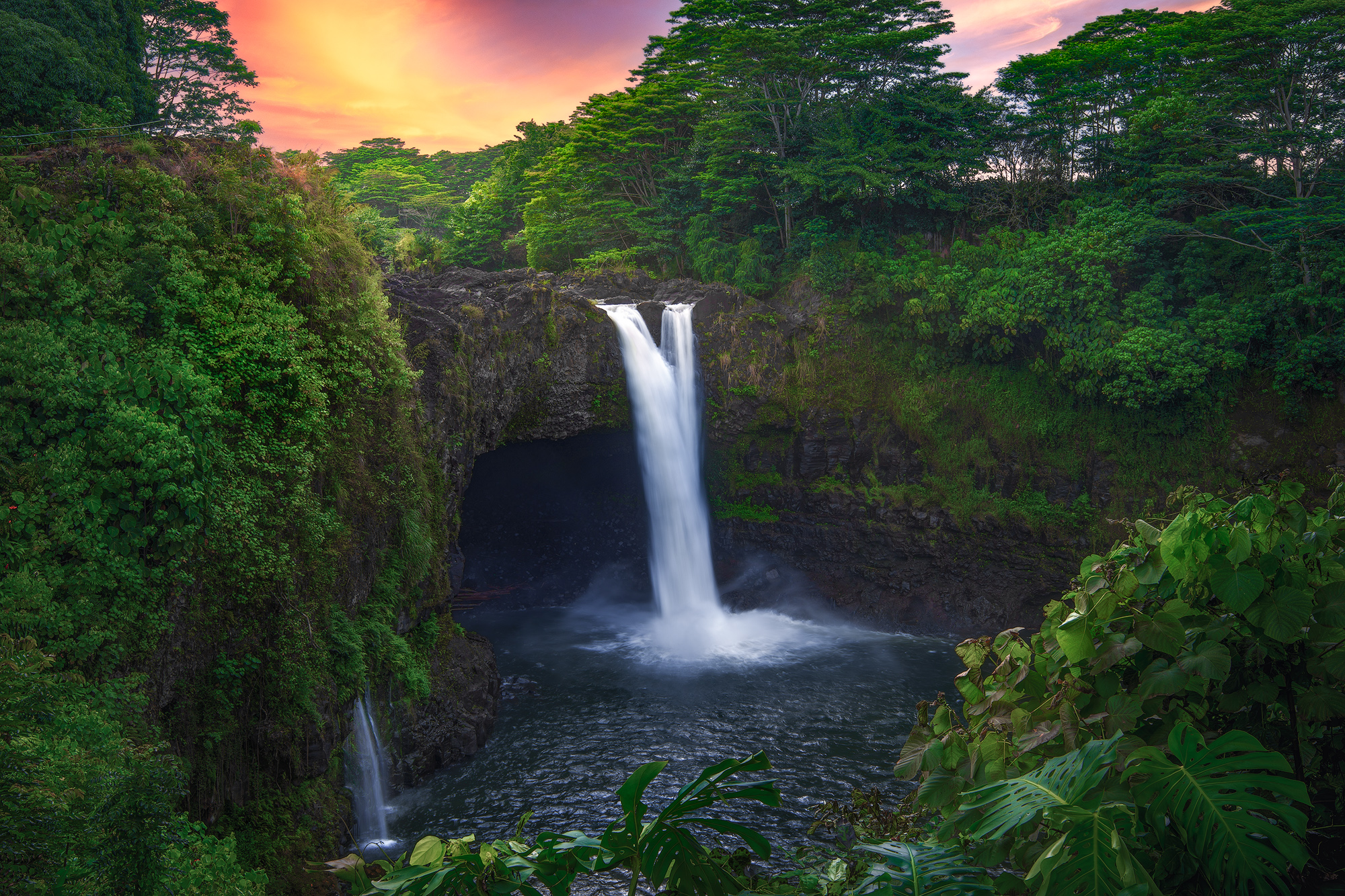 Twilight Over Waianuenue Rainbow Falls (Waianuenue) cascades into a cave basin in Hilo under a glowing orange and pink twilight sky surrounded by lush jungle.